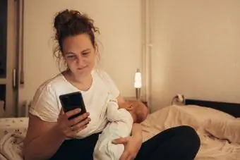 Mutter stillt ihr neugeborenes Kind zu Hause, während sie ein Smartphone benutzt