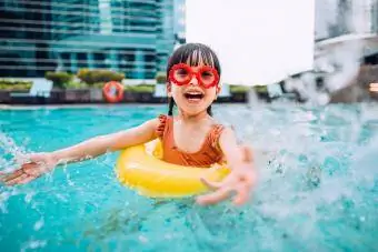 Niña juguetona con gafas de sol sonriendo alegremente, chapoteando y jugando alegremente con agua en la piscina durante las vacaciones de verano