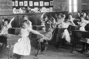 Djeca koja vježbaju gimnastiku dok sjede za svojim stolovima 1899