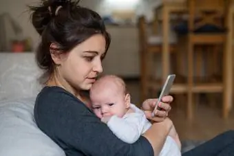 Mamă iubitoare și afectuoasă ținând copilul nou-născut în casă, folosind smartphone-ul.