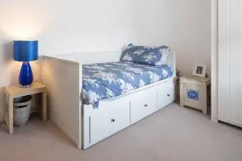 Pemandangan umum kamar tidur single dengan dinding putih dan tempat tidur bermotif bunga biru dengan mantel biru tergantung di belakang pintu di dalam rumah