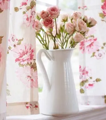 White enamelware pitcher na may maputlang pink na rosas sa windowsill na may mga vintage floral na kurtina