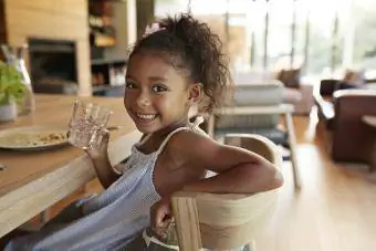 vajzë e ulur në tryezën e darkës me gotë ujë, duke buzëqeshur në kamerë