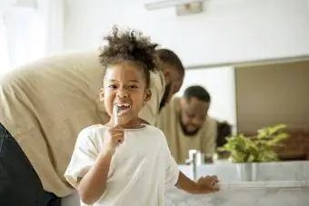 Bērns tīra zobus, kamēr viņas tēvs stāv aiz muguras vannas istabā