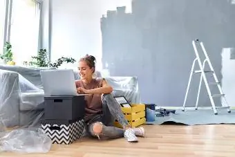 Wanita tersenyum bekerja pada komputer riba sambil duduk di tepi sofa di rumah
