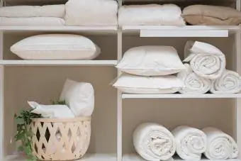Beyaz dolabın içinde yastık, yorgan gibi çeşitli ev eşyaları duruyor.