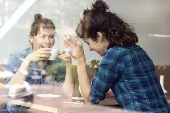 Duas mulheres rindo sentadas atrás da vidraça de uma cafeteria