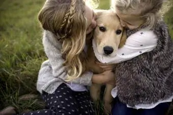 İki genç kız açık havada evcil köpeği kucaklıyor ve öpüyor.