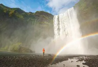 Mann blickt auf Wasserfall in idyllischer Landschaft und doppeltem Regenbogen