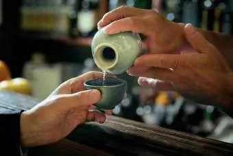 Primer plano de la mano del camarero vertiendo sake en un vaso que manos de un hombre adulto