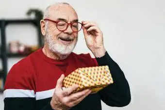 Pria senior tersenyum mengenakan kacamata melihat kotak hadiah di rumah