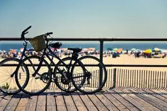 Sykler i silhuett, støttet ved kanten av strandpromenaden, mot et bakteppe av en overfylt strand om sommeren.