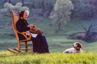 Une femme âgée est assise dehors sur une chaise à bascule épluchant des pommes