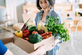 Gruaja merr një kuti plot me sende ushqimore organike shumëngjyrëshe dhe të freskëta