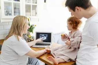 Nasmejana nosečnica, ki očetu na mizi v dnevni sobi prikazuje sliko ultrazvoka hčerki