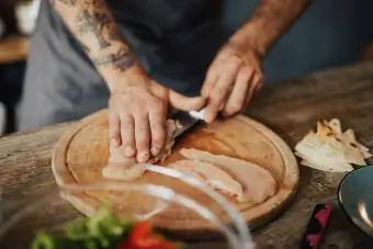Близък план на ръцете на главния готвач, които режат пилешко месо върху дървена дъска