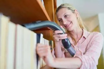 Mujer sonriente limpiando estantería con aspiradora en casa