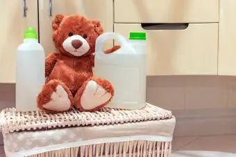 Um ursinho de pelúcia está sentado no cesto de roupa suja no banheiro ao lado de detergente e abrilhantador