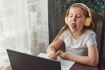 Pikkutyttö käyttää kannettavaa tietokonetta ja näyttää hampaansa ja hymynsä opiskellakseen kotona puheterapeutin kanssa