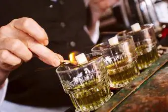 Cilvēka roka tur vieglus sērkociņu sildošus cukura kubiņus virs karotes uz absinta glāzēm pie bāra