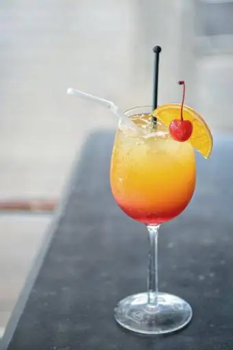 Cocktail roșu cu o felie de portocală și cireșe pe fundalul mesei