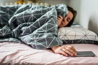 Wanita di tempat tidur melihat ponsel pintar