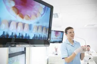 Tannlege med vernebriller og holder tannutstyr ved siden av skjermer som viser bilder av tenner