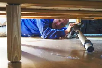 Bir adam bir apartman dairesinde yatağın altındaki tozu süpürüyor